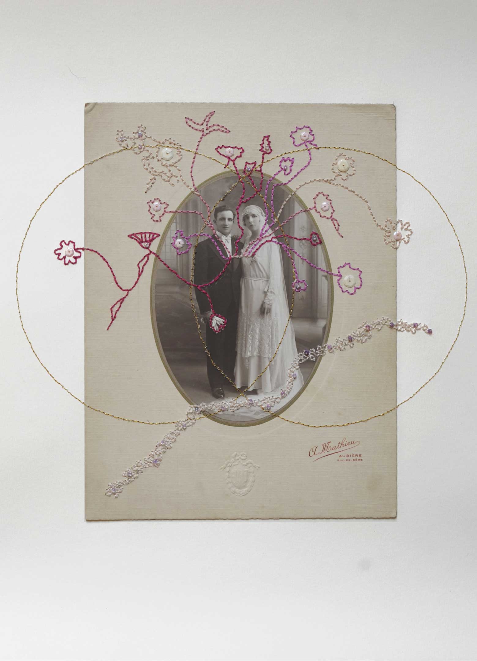 Photographie ancienne noir et blanc de mariage d'un couple au début du siècle, de forme ovale, entouré de broderies colorées et de 2 cercles brodés en fil d'or se croisant comme des alliances